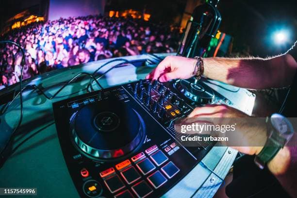 DJ's hands adjusting a mixing console overlooking a large, blurred crowd under vibrant lights.