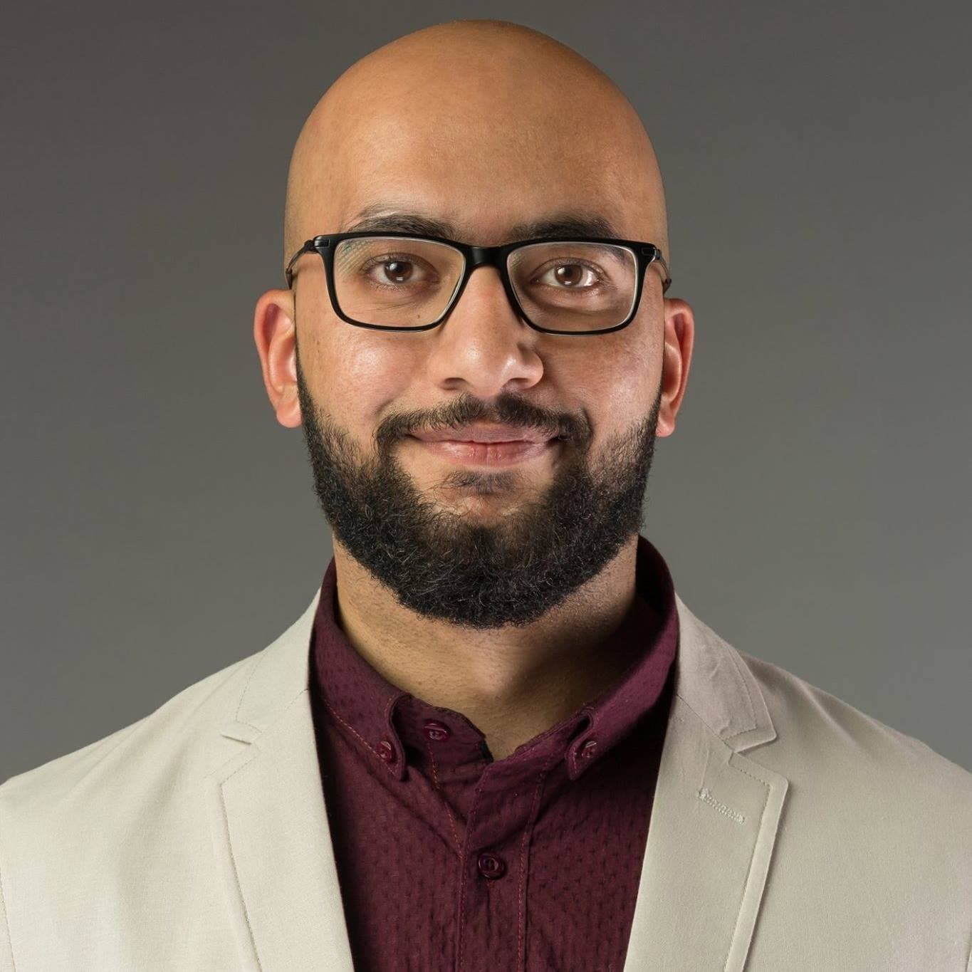 Bald man with a dark beard and glasses wearing a beige blazer over maroon shirt.